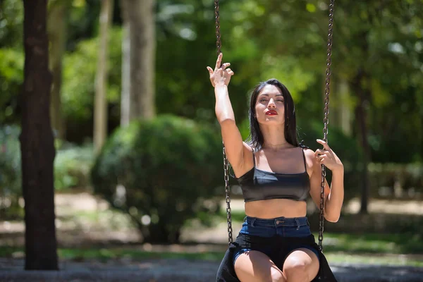 South American woman, young, pretty, brunette with leather top and short jeans, sitting on a swing in sensual and provocative attitude. Concept of beauty, diversity, sensuality and provocation.
