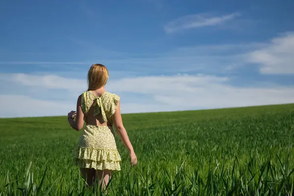 Young, beautiful, blonde woman in a yellow dress, walking through a green wheat field, seen from the back, alone, calm and peaceful. Concept beauty, relaxation, tranquility, calm, fields, meadows.
