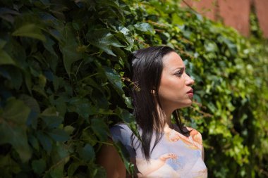 Genç bir kadın, güzel, esmer ve baskılı gömlekli, yeşil sarmaşık arka planında sakin ve rahat, güneş ışınlarını alan bir Güney Amerikalı. Güzelliği, modayı, bitkileri kavra.