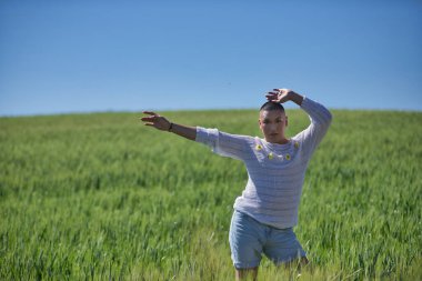 Genç, Güney Amerikalı, ikili olmayan, ağır makyajlı, yeşil buğday tarlasının ortasında, doğal papatyalarla beyaz bir kazakla poz veren, kolları havada. Kavram kraliçesi, lgbtq +, gurur, nonoş.