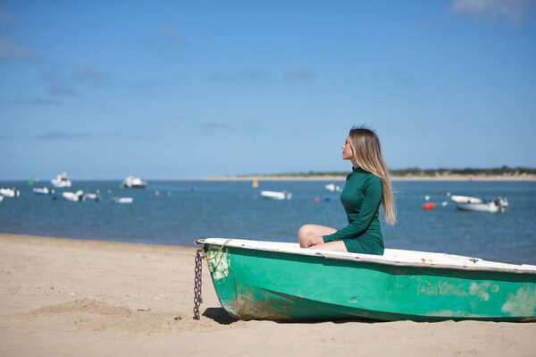 Young, beautiful, blonde, green-eyed, young woman in a green dress, sitting in a boat, looking at infinity, relaxed and calm, on the beach, with the sea in the background. Concept vacation, travel.