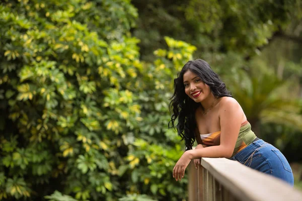 South American woman, young, beautiful, brunette, with colorful top and jeans leaning on a wooden railing looking at the camera smiling and happy. Concept of beauty, trend, ethnicity, diversity.