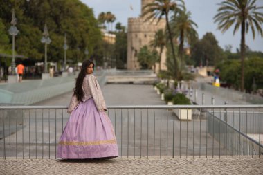 Genç bir kadın, İspanyol, güzel, esmer, şık bir menekşe elbise giymiş, şehri ziyaret ederken korkuluklara yaslanmış poz veriyor. Güzellik kavramı, moda, trend, zarafet, antika.