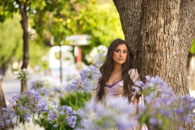 Genç, İspanyol, güzel, esmer, zarif bir menekşe elbise giymiş, bir ağacın gövdesine yaslanmış, menekşe çiçeklerinin arasından görülüyor. Güzellik kavramı, moda, trend, zarafet, bahar.