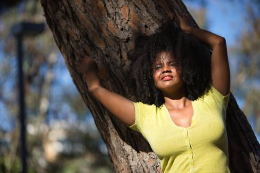 Genç bir kadın, güzel ve siyah, afro saçlı, sarı gömlekli, şehvetli ve kışkırtıcı bir tavırla bir ağacın gövdesine yaslanmış. Duygusallığı, provokasyonu, flörtü kabul et..