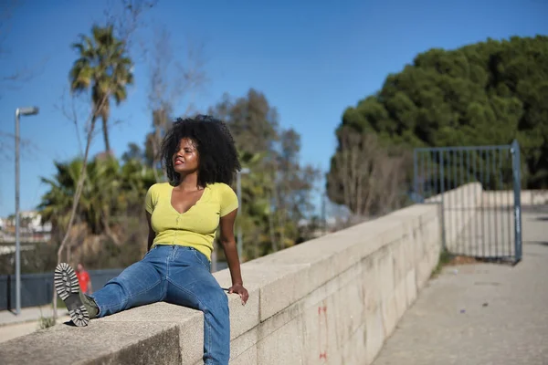 Young, beautiful, black woman with afro hair, wearing a yellow t-shirt, jeans and boots, sitting on a stone wall, relaxed and calm, sunbathing. Concept of relaxation, tranquility, peace.