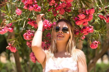 Güney Amerikalı bir kadının portresi, genç, güzel ve güneş gözlüklü, gülümseyen ve mutlu bir şekilde pembe çiçeklerin yanında duran bir sarışın. Kavram eğilimi, güzellik, moda.