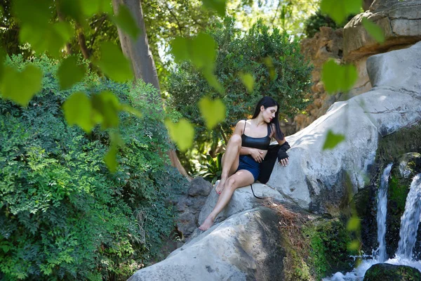 South American woman, young, pretty, brunette wearing leather top and short jeans, sitting on a large stone while watching a waterfall. Concept of beauty, diversity, water, nature.