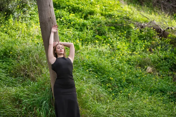 Genç bir kadın, sarışın ve güzel, siyah elbiseli, bir ağaç gövdesine yaslanmış, doğanın ortasında, bağlı ve sevecen. Doğa, huzur, mutluluk, sükunet, ağaçlar.