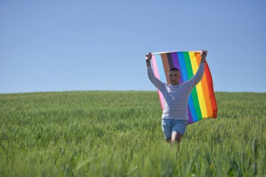 İkili olmayan biri, genç ve Güney Amerikalı, çok makyajlı, yeşil buğday tarlasının ortasında eşcinsel bir gurur bayrağı sallıyor. Konseptin kraliçesi, lgbtq +, gurur, nonoş.