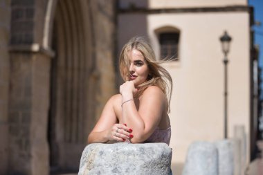 Genç bir kadının portresi, sarışın, yeşil gözlü, pembe bluzlu, taş bir sütuna yaslanmış, rüzgarın saçlarını hareket ettirmesine izin veren. Güzelliği kavra, saç stillerini, rüzgarı, yeleyi.