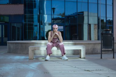 Genç, çekici, eşcinsel bir adam, ağır makyajlı, pembe saçlı, üst ve kot pantolonlu, dalgın, elleri çenesinde dinleniyor, taş bir bankta oturuyor, güneş ışınlarını yakalıyor. Kavram lgtbiq +, gay, gurur, makyaj.