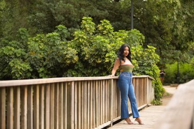 Güney Amerikalı bir kadın, genç, güzel, esmer, renkli bluzlu ve kot pantolonlu ahşap korkuluklara yaslanmış gülümseyen ve mutlu bir şekilde kameraya bakıyor. Güzellik kavramı, eğilim, etnik köken, çeşitlilik.