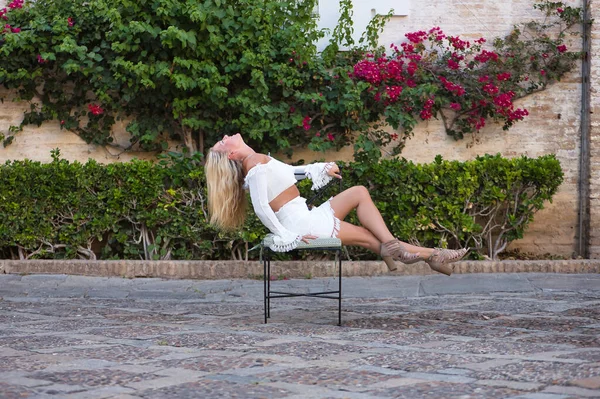 Young, beautiful, blonde American woman in a white dress, with her head back and her hair falling down sitting on a chair in the street, during her trip to a European city. Concept travel, tourism,