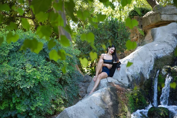 South American woman, young, pretty, brunette in leather top and short jeans, sitting on a large stone looking at the camera next to a waterfall. Concept of beauty, diversity, water, nature.