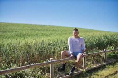 İkili olmayan, genç ve Güney Amerikalı, ağır makyajlı, yeşil buğday tarlasının yanında, ahşap bir çitin üzerinde kameraya bakıyor. Kavram kraliçesi, lgbtq +, gurur, nonoş.