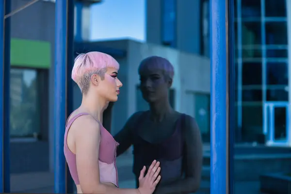 stock image Young, attractive, gay man, heavily makeup, with pink hair and top, looking at a glass wall in which he sees himself reflected, looking for himself. Concept lgtbiq+, gay, pride, makeup, fashion, trend