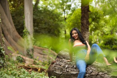 Güney Amerikalı bir kadın, genç, güzel, esmer, renkli bluzlu ve kot pantolonlu, bir ağaç gövdesinde oturuyor, gülümsüyor, mutlu ve rahat. Güzellik, moda, trend, etnik köken, çeşitlilik kavramı.