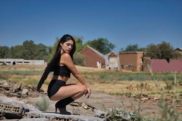 South American woman, young, beautiful, brunette with lingerie and black wings, posing crouching receiving sun rays in the middle of the ruins of a building. Concept angels, beauty, costumes, wings.