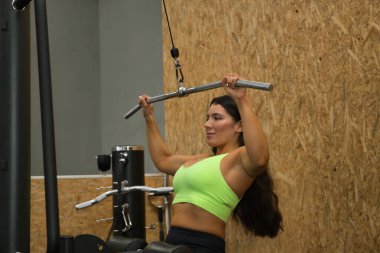 Genç, güzel, esmer kadın, sarı bluzlu ve siyah taytlı, spor salonunda sırt egzersizleri için göğüs barfiksi çekiyor. Fitness konsepti, jimnastik, vücut geliştirme, trend.