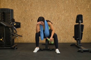 Genç, güzel, esmer, sarı bluzlu, siyah taytlı, boynunda havluyla yorgun, bitkin, bunalımda ve hayal kırıklığına uğramış spor salonundaki bir bankta oturan bir kadın. Fitness konsepti, jimnastik.