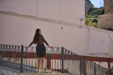 Genç, güzel, zarif siyah elbiseli esmer kadın, arkası dönük, güzel beyaz bir Endülüs köyünde parmaklıklara yaslanmış kameraya bakıyor. Güzelliği, modayı, eğilimi kavra.