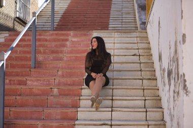 Endülüs 'ün güzel beyaz bir köyünün merdivenlerinde oturan zarif, siyah elbiseli, dikkati dağınık bakışlı genç, güzel, esmer kadın. Güzellik, moda, trend, seyahat, kırsal.