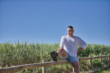 İkili olmayan, genç ve Güney Amerikalı, ağır makyajlı, yeşil buğday tarlasının yanında, ahşap bir çitin üzerinde kameraya bakıyor. Kavram kraliçesi, lgbtq +, gurur, nonoş.