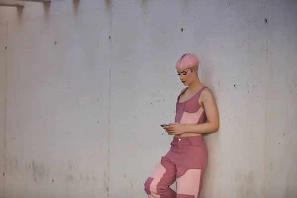 Stock image Young, attractive, gay man, heavily makeup, with pink hair, top and jeans, checking his mobile phone, leaning against a wall. LGTBIQ+ concept, gay, pride, makeup, fashion, trend.