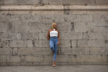 Olgun bir kadın, güzel, sarışın, beyaz bluzlu ve kot pantolonlu, gri bir duvara yaslanmış, yalnız, bağımsız ve güçlü. Güzellik, moda, trend, seyahat, olgunluk kavramı.
