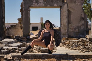 Güney Amerikalı bir kadın, genç, güzel, esmer, iç çamaşırı ve siyah kanatları olan, yıkılmış binaların ortasına çömelmiş, rahat ve sakin bir şekilde poz veren. Melekleri, güzelliği, kostümleri, kanatları, cadılar bayramını.