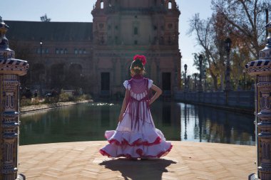 Flamenko yapan tipik flamenko elbiseli bir kız, sırtı dönük, Seville 'de güzel bir meydanda, bir gölün yanında güneş ışınları alan. Konsept dansı, flamenko, tipik İspanyol, İspanya.