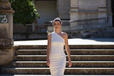 Beyaz takım elbiseli, genç, güzel, sarışın bir kadın merdivenin yanında mutlu ve gülümseyen kendinden emin bir şekilde poz veriyor. Güzellik, moda, moda, model konsepti.