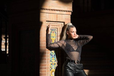 Genç, güzel, sarışın, mavi gözlü, şeffaf siyah gömlekli, deri pantolonlu, duvara yaslanmış, karanlıkta güneş ışığı alan bir kadın..