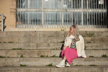 Genç, güzel, sarışın kadın, mavi gözlü, beyaz püsküllü ceketli ve pembe etekli, eli çenesinde, sonsuzluğa bakıyor, rahat ve sakin bir şekilde bazı basamaklarda oturuyor..