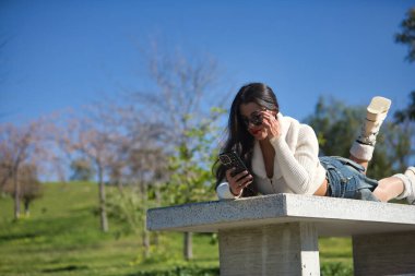 Genç, güzel, beyaz kazaklı, kot etekli ve güneş gözlüklü esmer kadın güneşli bir günde parktaki bankta uzanan akıllı telefonuyla sosyal medyayı kontrol ediyor. Güzellik, moda, moda konsepti.