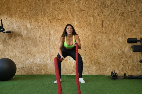 stock image Young, beautiful, brunette woman in yellow top and black leggings, doing rope battle exercises in gym. Fitness concept, gymnastics, bodybuilding, trend.