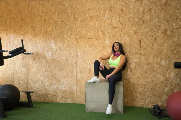 stock image Young, pretty, brunette woman, pink headphones, wearing yellow top and black leggings, sitting on a wooden jumping box, relaxed and calm in the gym. Fitness concept, gymnastics, bodybuilding, trend.