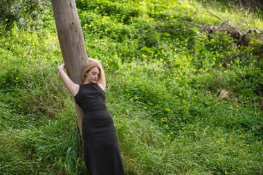 Genç bir kadın, sarışın ve güzel, siyah elbiseli, bir ağaç gövdesine yaslanmış, doğanın ortasında, bağlı ve sevecen. Doğa, huzur, mutluluk, sükunet, ağaçlar.