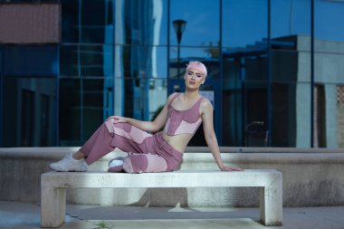 Genç, çekici, eşcinsel, makyajlı, pembe saçlı, bluzlu, kot pantolonlu, kameraya bakan, taş bir bankta oturan, güneş ışığı alan bir adam. Kavram lgtbiq +, gay, gurur, makyaj, moda, trend.