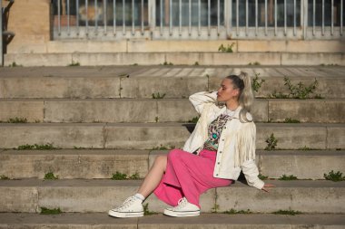Genç, güzel, sarışın, gözleri kapalı, beyaz püsküllü ceket ve pembe etekli, rahat ve sakin bir şekilde merdivenlerde oturuyor. Güzellik kavramı, moda, milenyum, trend.