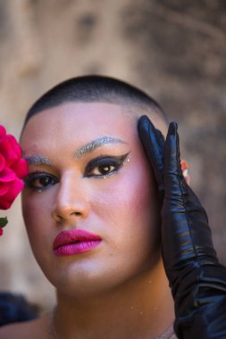 İkili olmayan, genç ve Güney Amerikalı, makyajlı, siyah kanatlı, deri eldivenli, korseli, yüzünde güllerle poz veren birinin yakın plan portresi. lgbtq + kavramı. Dikey konum.