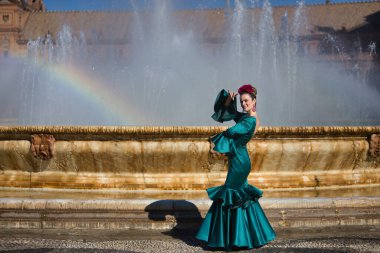 Genç, güzel, tipik yeşil flamenko takım elbiseli sarışın kadın, suda yansıyan gökkuşağı ile bir çeşmenin yanında dans ediyor. Flamenko konsepti, tipik İspanyolca, Sevilla, Endülüs.