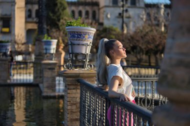 Genç, güzel, sarışın bir kadın, gözleri kapalı ve yazılı bir tişörtü var, korkuluklara yaslanmış, güneşlenen, rahat ve sakin. Güzellik kavramı, moda, milenyum, trend.