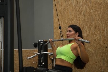 Genç, güzel, esmer kadın, sarı bluzlu ve siyah taytlı, spor salonunda sırt egzersizleri için göğüs barfiksi çekiyor. Fitness konsepti, jimnastik, vücut geliştirme, trend.