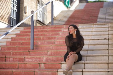 Endülüs 'ün güzel beyaz bir köyünün merdivenlerinde oturan zarif, siyah elbiseli, dikkati dağınık bakışlı genç, güzel, esmer kadın. Güzellik, moda, trend, seyahat, kırsal.