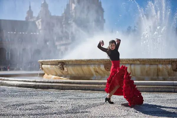 Genç, güzel, esmer, siyah tişörtlü ve kırmızı etekli kadın Sevilla 'daki güzel bir çeşmenin önünde flamenko dansı yapıyor. Flamenko konsepti, dans, sanat, tipik İspanyolca.