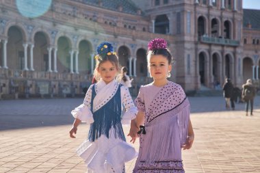 Flamenko dansı yapan iki kız kameraya bakıyor, el ele tutuşuyorlar, Sevilla 'da güzel bir meydanda tipik flamenko elbiseler giyiyorlar. Dans konsepti, flamenko, tipik İspanyolca, Sevilla, İspanya.