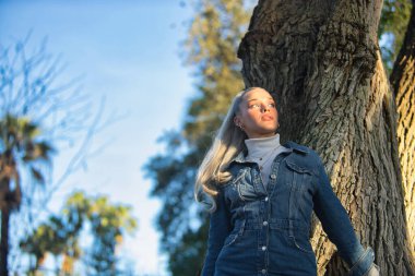 Kadın, genç, güzel, sarışın, mavi gözlü ve kot elbiseli, aşağıdan görünen, güneş ışınlarını gün batımında alan büyük bir ağacın gövdesine yaslanan sonsuzluğa bakıyor..