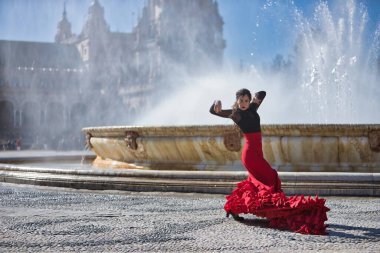 Genç, güzel, esmer, siyah tişörtlü ve kırmızı etekli kadın Sevilla 'daki güzel bir çeşmenin önünde flamenko dansı yapıyor. Flamenko konsepti, dans, sanat, tipik İspanyolca.